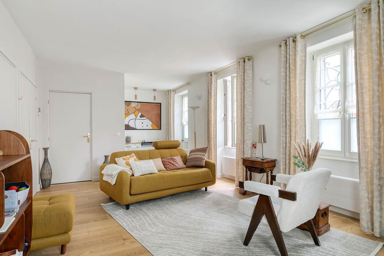 Le cabinet d'Emmanuelle Galy à Paris 14e : une pièce lumineuse aux grandes fenêtres avec un canapé moutarde, un fauteuil blanc et un parquet de chêne clair.